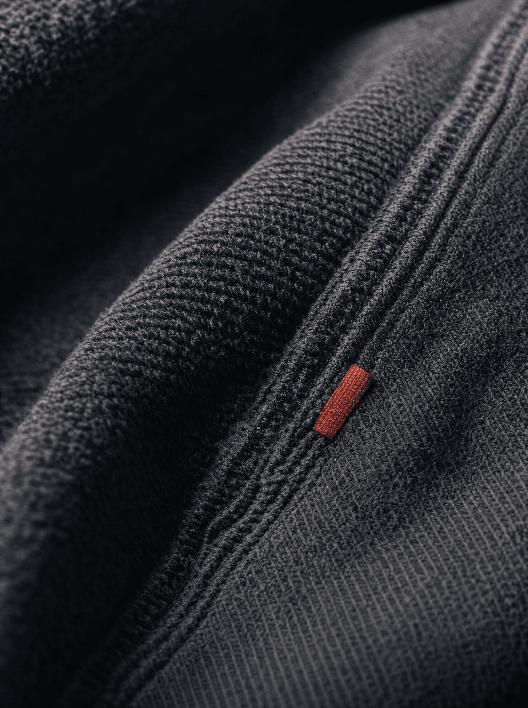 Extreme macro of the Vantor 14oz cotton-fleece loopback seam under raking side light, single brick-red care tab in frame