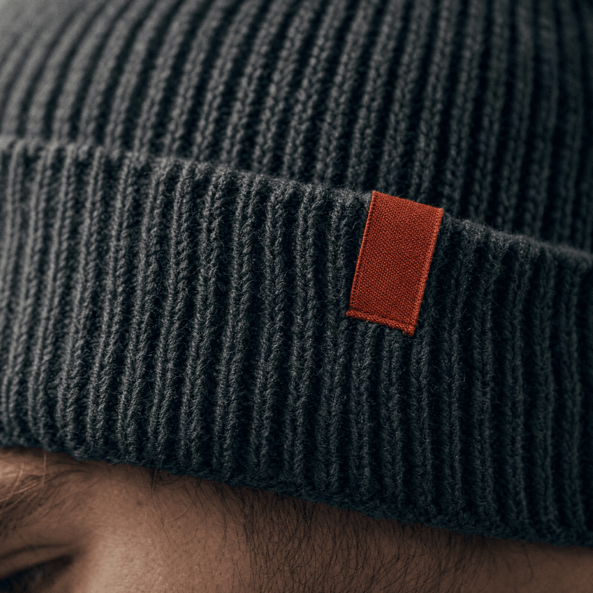Close-up of the ribbed charcoal beanie cuff with the brick-red Vantor accent tab, raking side light
